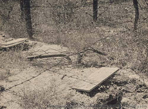 [A fougasse flame thrower emplaced under a barbed-wire entanglement. Only the nozzle and propellant cylinder show above ground.]